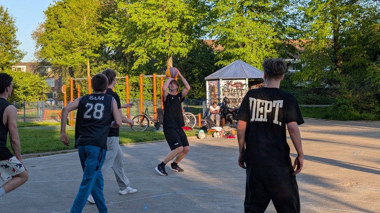 Jongeren spelen basketbal op een buitenveld omringd door bomen, met een jongen die een schot probeert te maken.