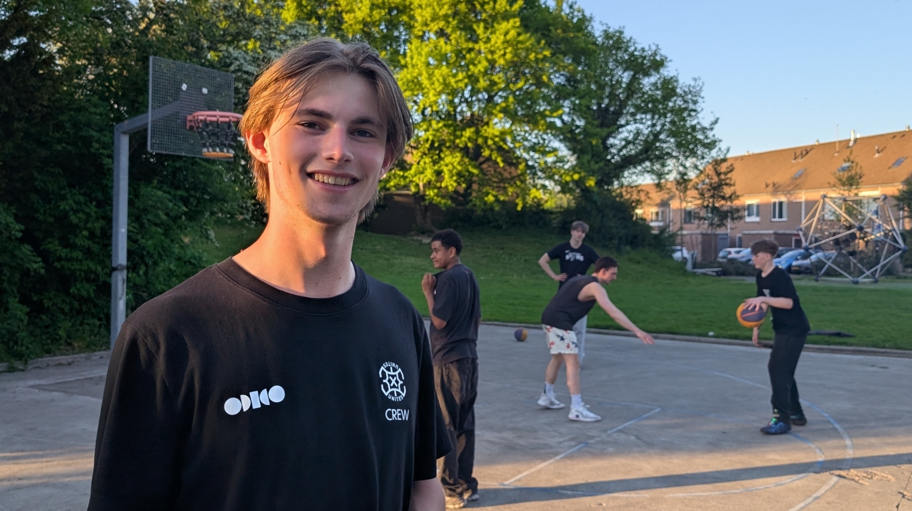 Jongeman in een zwart T-shirt glimlacht op een basketbalveld; op de achtergrond spelen anderen basketbal.