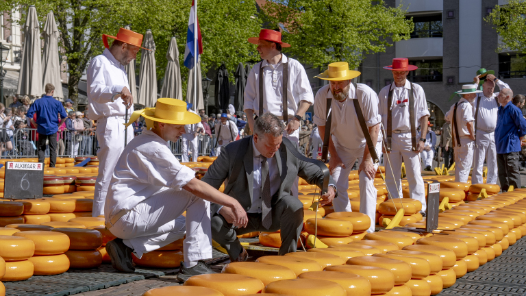 Mannen in traditionele kleding inspecteren kaas tijdens een kaasmarkt, omringd door stapels kazen.