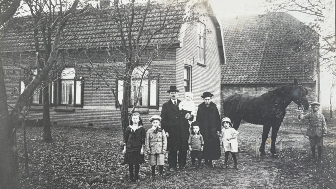 Oude zwart-witfoto van een gezin naast een huis met een paard, ingelijst aan een muur.