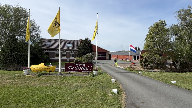 Ingang van Camping 'De Boekel' met vlaggen, een grote gele klomp en een oprijlaan, omringd door groen en gebouwen.