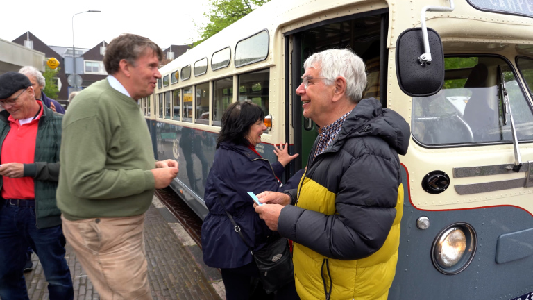 Mensen stappen lachend in een ouderwetse bus; een man toont zijn kaartje.