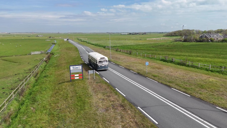 Een oude bus rijdt over een rustige weg door een landelijk gebied met groene velden en een paar windmolens in de verte.