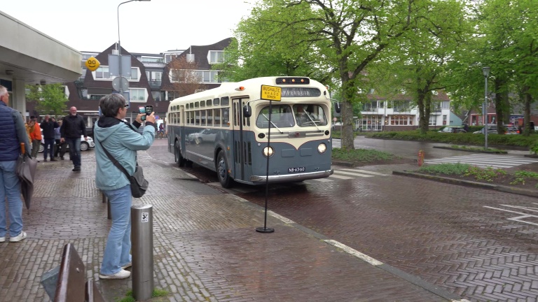 Een klassieke bus rijdt over een natte straat, terwijl mensen foto's maken en toekijken.