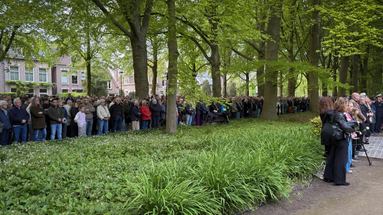 Grote groep mensen verzameld in een parkachtig gebied met bomen, blijkbaar bij een plechtige gebeurtenis.