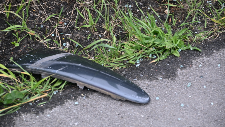 Zwart stuk autolak ligt op de grond naast gras en asfalt, omringd door kleine glasscherven.