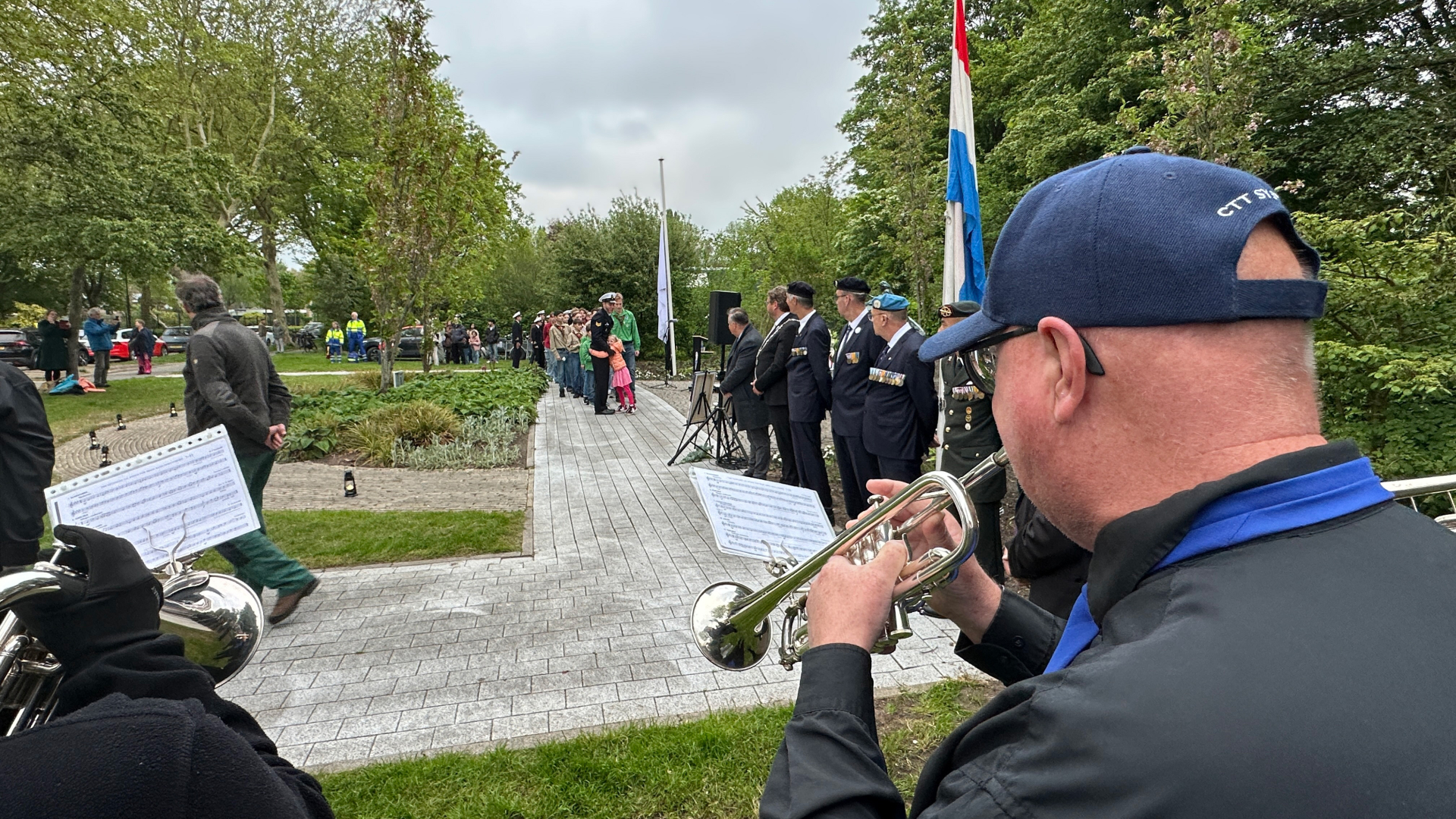 Muzikanten spelen trompetten tijdens een ceremonie, met een rij mensen en veteranen bij een vlag in een parkachtige omgeving.