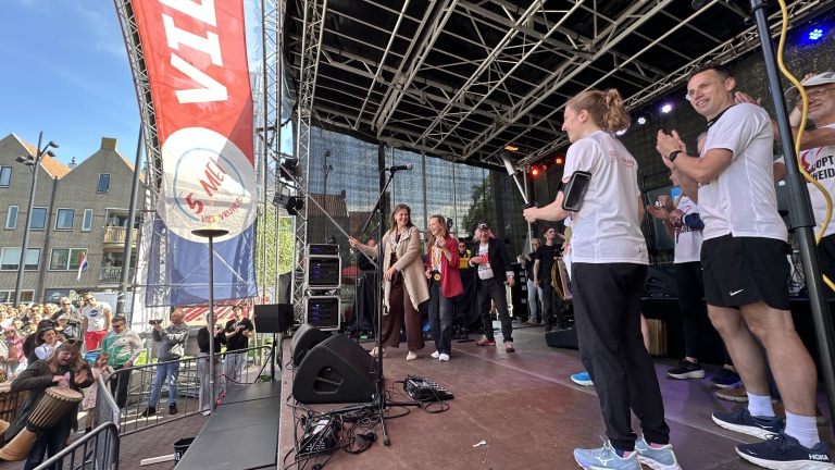 Mensen op een podium tijdens een evenement met een "5 mei" vlag op de achtergrond en toeschouwers op de voorgrond.