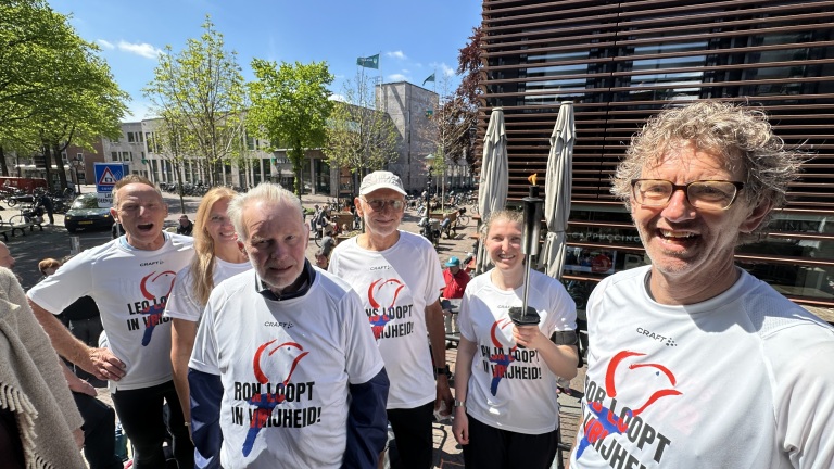 Een groep mensen in witte T-shirts met teksten verzameld buiten in de zon, met een vrouw die een fakkel vasthoudt.