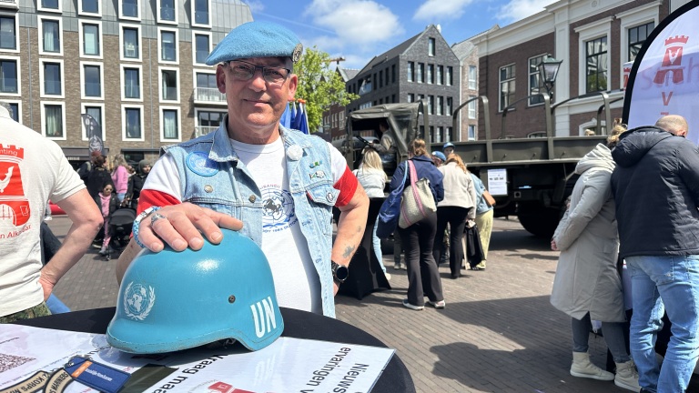 Man met blauwe UN-helm en baret staat buiten met een informatiestand, omringd door mensen en gebouwen.