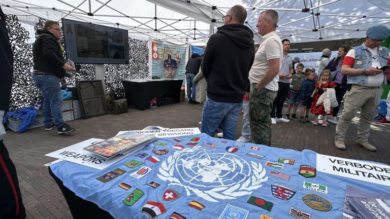Een groep mensen verzamelt zich in een tent bij een presentatie met vlaggen van de VN en diverse landen op tafel. Posters met "Verboden Toegang" zijn zichtbaar.