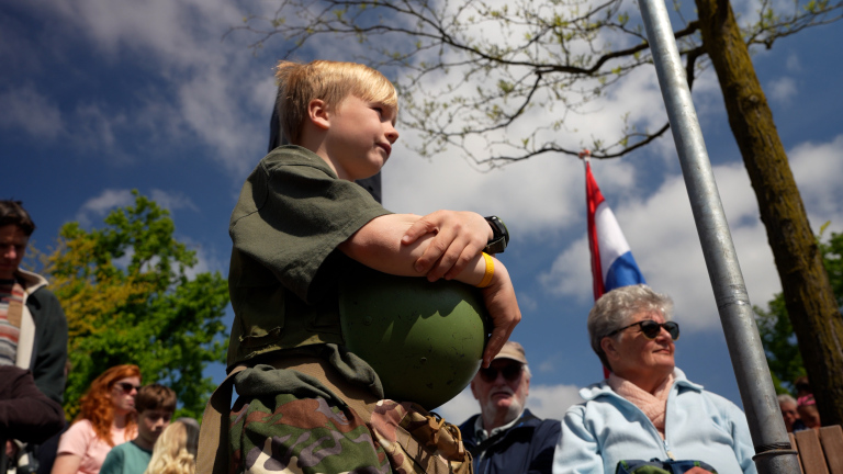 Jongen in legeroutfit houdt een helm vast, omringd door mensen, met een Nederlandse vlag op de achtergrond.