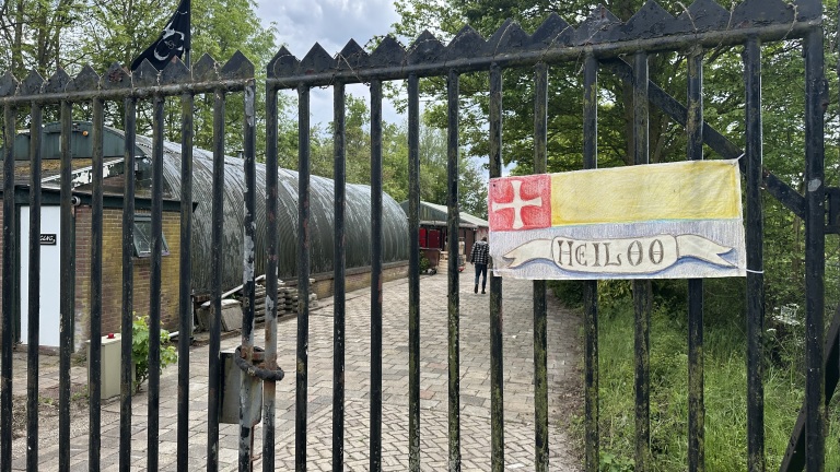 Het hek van een terrein met een gebouw op de achtergrond, en een bord met "Heiloo" hangt aan het hek.