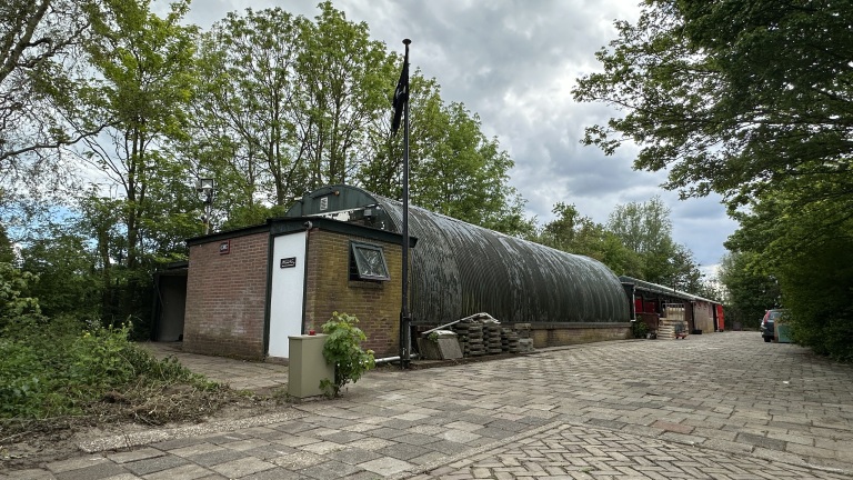 Bakstenen gebouw met metalen dak in een groene omgeving, met opgestapelde materialen ernaast en een geplaveide oprit.