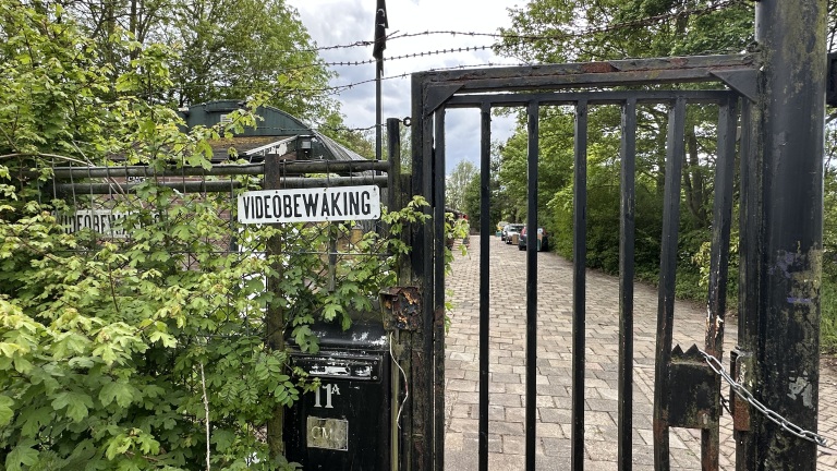 Openstaande ijzeren poort met bord "videobewaking", omgeven door bladeren, leidt naar een weg met geparkeerde auto's.