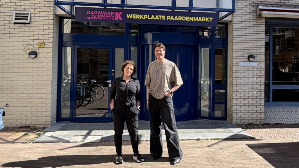 Twee mensen staan voor een gebouw met het bord "Werkplaats Paardenmarkt" en twee kleurrijke vlaggen.