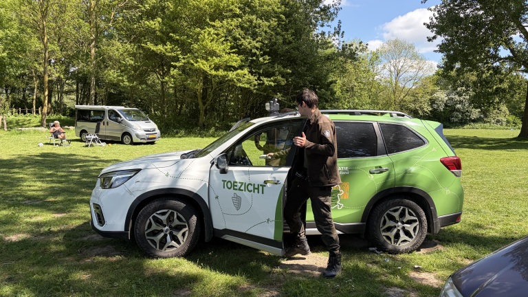 Man stapt uit een groen-witte toezichtauto in een park, met een zilveren busje en een persoon op stoelen op de achtergrond.