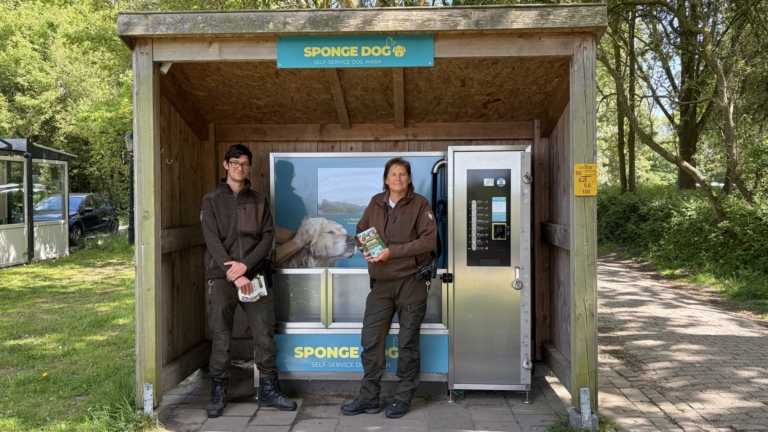 Twee personen staan voor een zelfbedienings-washok voor honden met het bordje "Sponge Dog", omgeven door een groene omgeving.