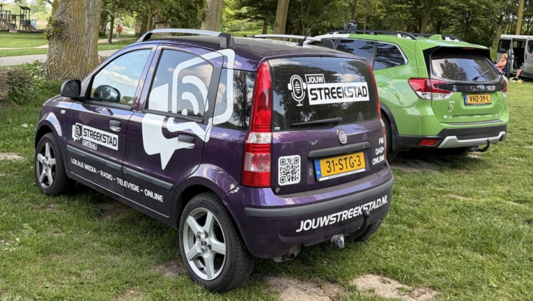 Paarse auto met logo van "Jouw Streekstad" en bijbehorende tekst, geparkeerd naast groene auto op grasveld met bomen.