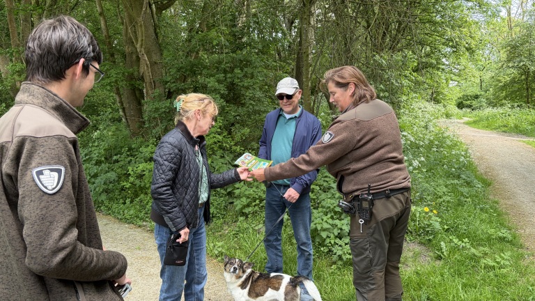 Vier mensen staan in een bos, waarbij een vrouw een folder ontvangt van een man in een groen uniform. Er is een hond aanwezig.