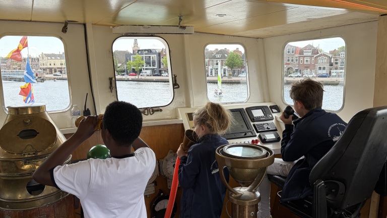 Kinderen op een schip zijn bezig met navigeren; een kijkt door een verrekijker, een ander praat in een microfoon.