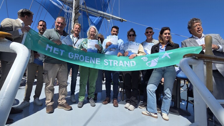 Groep mensen op een schip houdt een groen spandoek met "Het Groene Strand" vast, onder een blauwe lucht.