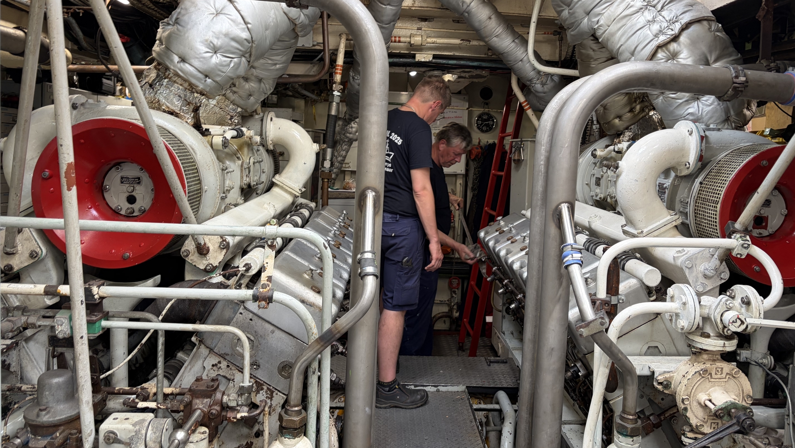 Twee mensen werken in een machinekamer met grote motoronderdelen en ingewikkelde leidingen.