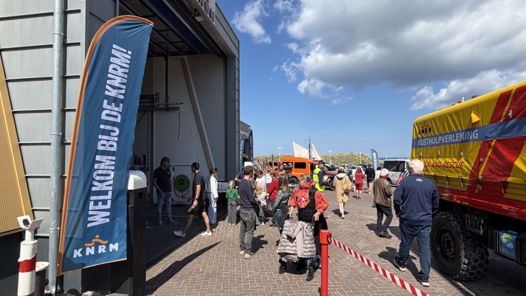 Mensen staan in de rij bij een KNRM-gebouw met een spandoek dat zegt "Welkom bij de KNRM"; een voertuig voor kusthulpverlening is zichtbaar.