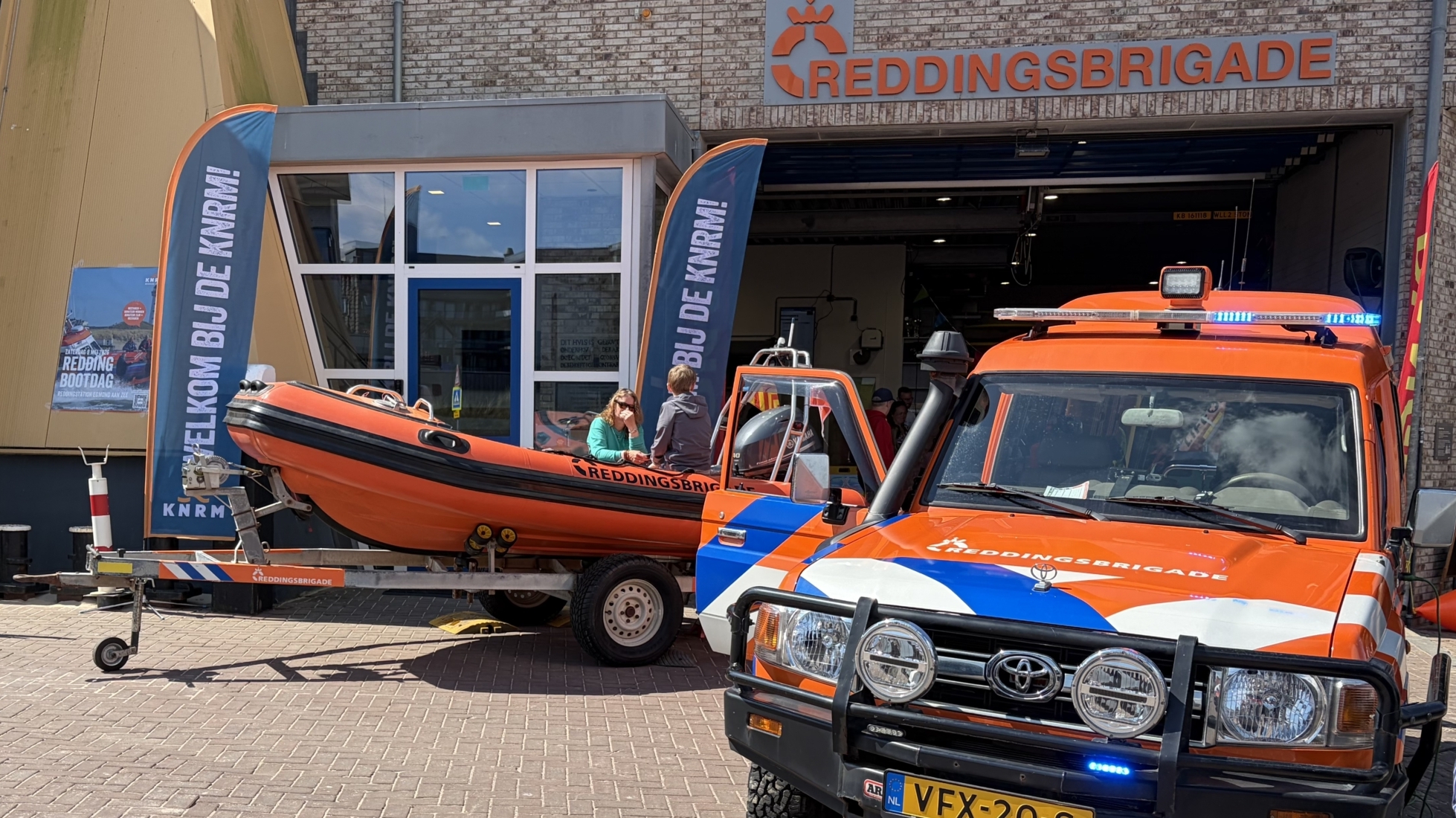 Reddingsbrigade voertuig en reddingsboot staan voor een gebouw, met mensen en een zwarte hond nabij.