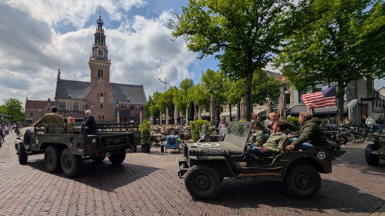 Militaire voertuigen en personen in legerkleding rijden door een plein met een historische kerk op de achtergrond.