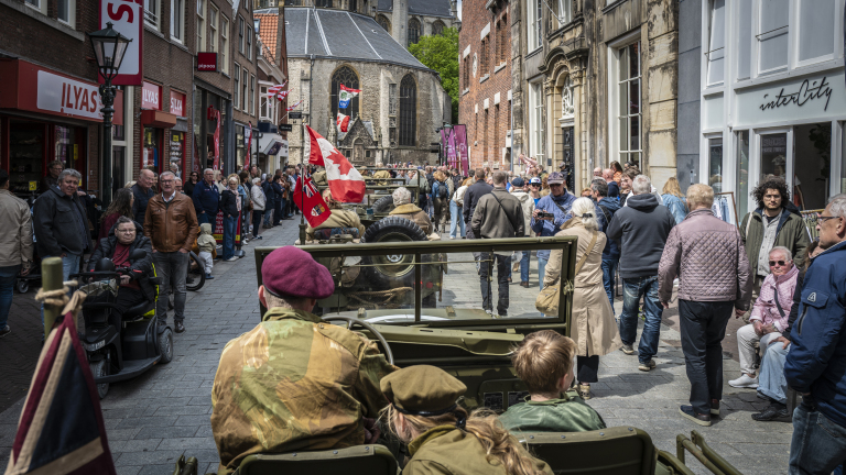 Een straat gevuld met toeschouwers en militaire voertuigen met vlaggen, tegen de achtergrond van historische gebouwen.
