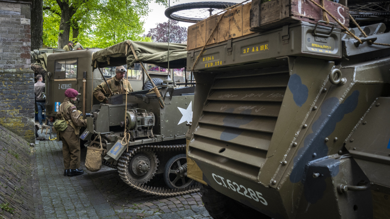 Mensen in militaire uniformen staan bij historische voertuigen met kisten en uitrusting op een geplaveide straat.