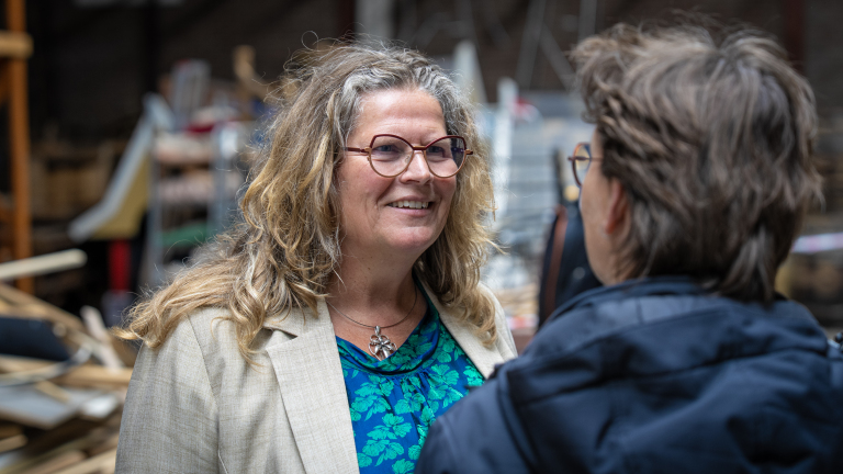 Twee personen in gesprek, een vrouw met bril en krullend blond haar glimlacht, terwijl ze een bloemrijke blouse draagt.