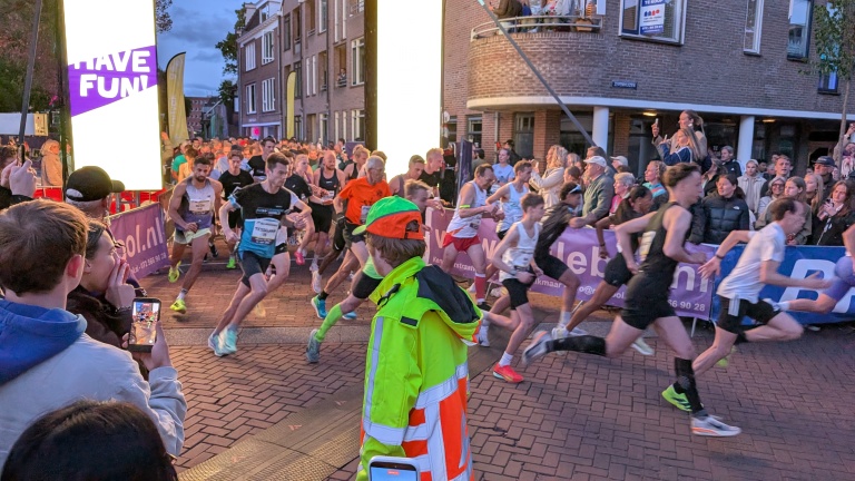 Hardlopers starten een race in een drukke straat, omgeven door toeschouwers en een verkeersregelaar in een felgekleurd vest.