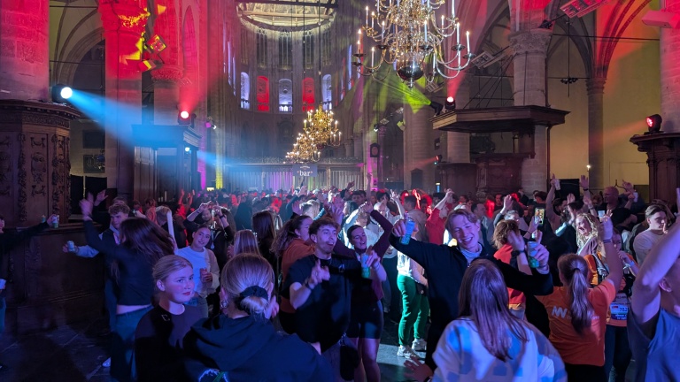Mensen dansen en feesten in een verlichte kerk met kleurrijke lichten en kroonluchters aan het plafond.