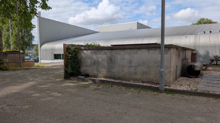 Industrieel gebouw met een gebogen stalen dak en betonnen muren, omgeven door een bestraat terrein met enkele planten.