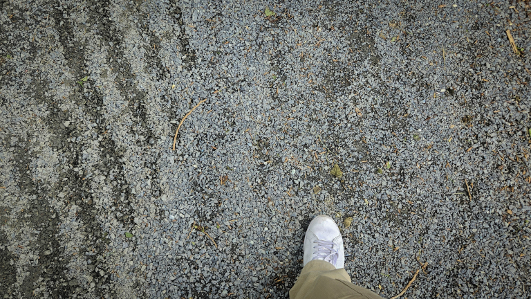 Een voet met witte sneaker op een grindpad vol kleine steentjes en sporen.