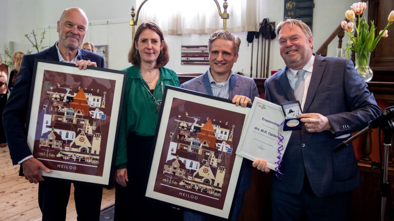Vier mensen poseren met ingelijste posters van Heiloo en een oorkonde met een medaille in een ceremonieruimte.