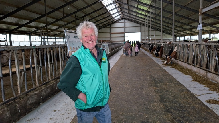 Man in groene jas staat glimlachend in een koeienstal, met kinderen en volwassenen op de achtergrond.