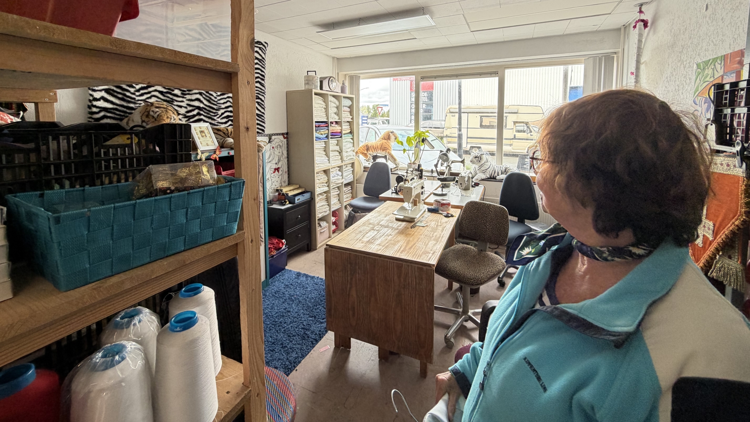Een vrouw in een blauwe jas kijkt rond in een naaiatelier met planken, garenrollen en een tafel vol naaimachines.
