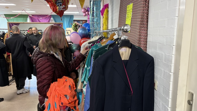 Een vrouw bekijkt kleding aan een rek in een drukke tweedehandswinkel, met feestelijke ballonnen op de achtergrond.