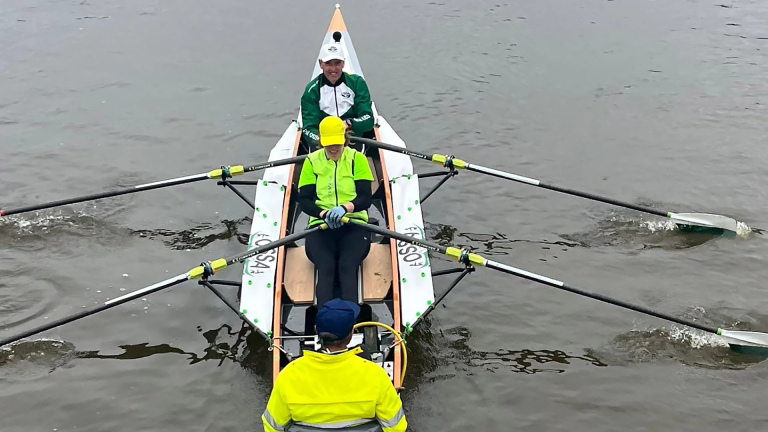 Twee mensen roeien in een dubbele boot met een stuurman op het water, gekleed in felgekleurde kleding.