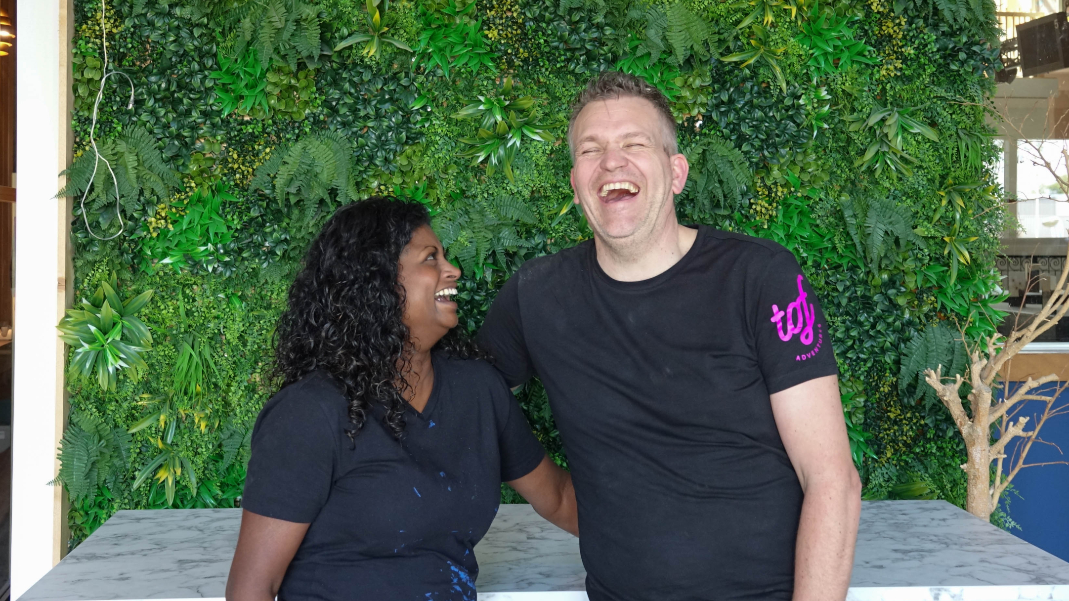Twee mensen lachen samen voor een groene plantenwand.