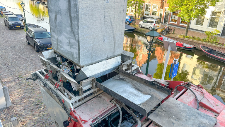 Vrachtwagen met open laadbak tilt afvalcontainer in een stedelijk gebied, naast een gracht met geparkeerde auto's en boten.