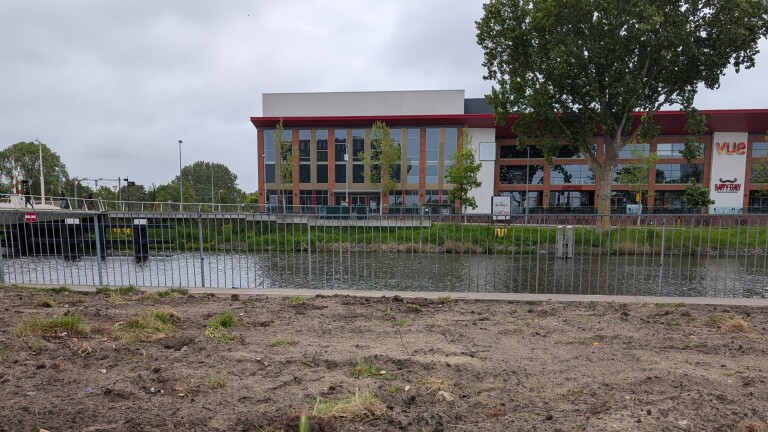 Gebouw met grote ramen aan de overkant van een gracht, met bomen en een brug aan de linkerkant.