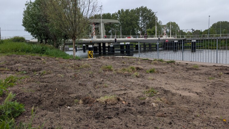 Een zandige oever met gras en enkele bomen, naast een open brug over een waterweg op een bewolkte dag.