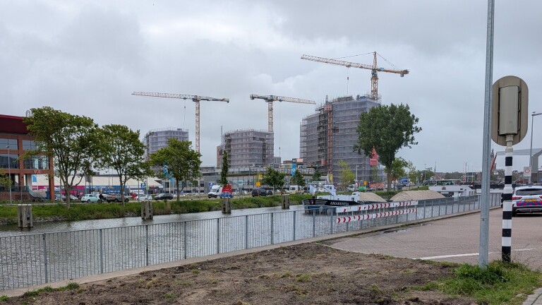 Bouwkranen en gebouwen in opbouw langs een waterweg, met auto's geparkeerd en een schip genaamd "Amarenta" op het kanaal.