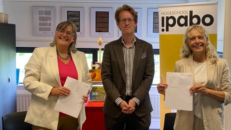 Drie mensen poseren in een kantoor bij een banner met de tekst “Hogeschool Ipabo”, twee van hen houden getekende documenten vast.