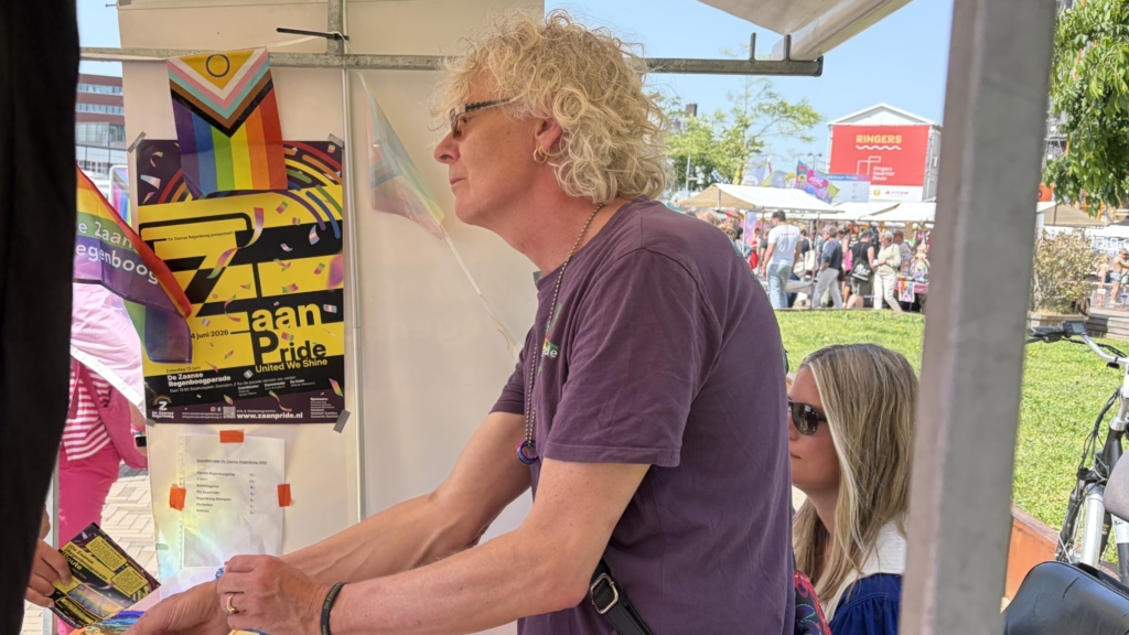 Een persoon met krullend haar staat achter een kraam met kleurrijke folders tijdens een buitenfestival.