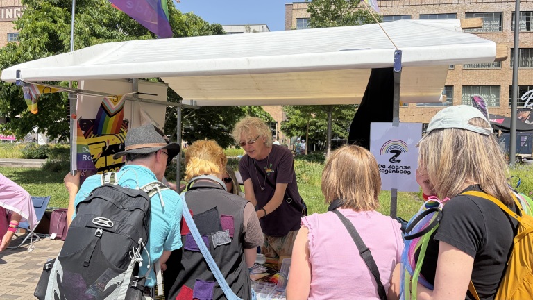 Een groep mensen verzamelt zich bij een kraam met de tekst "De Zaanse Regenboog" tijdens een zonnig buitenfestival.